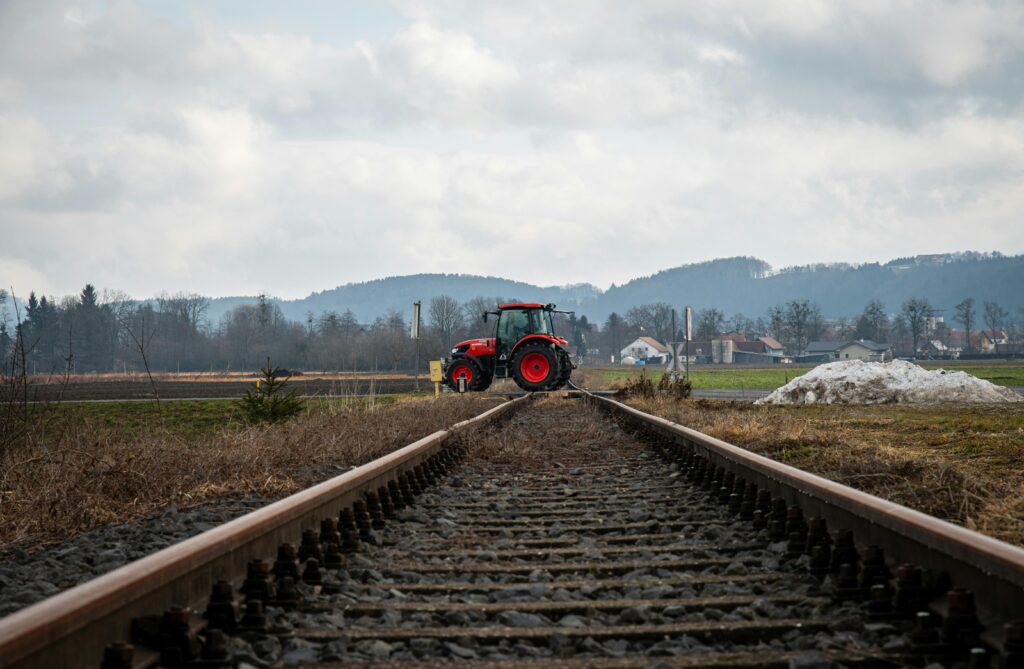A red tractor crossing railway tracks in a rural area with hills and a cloudy sky backdrop.
